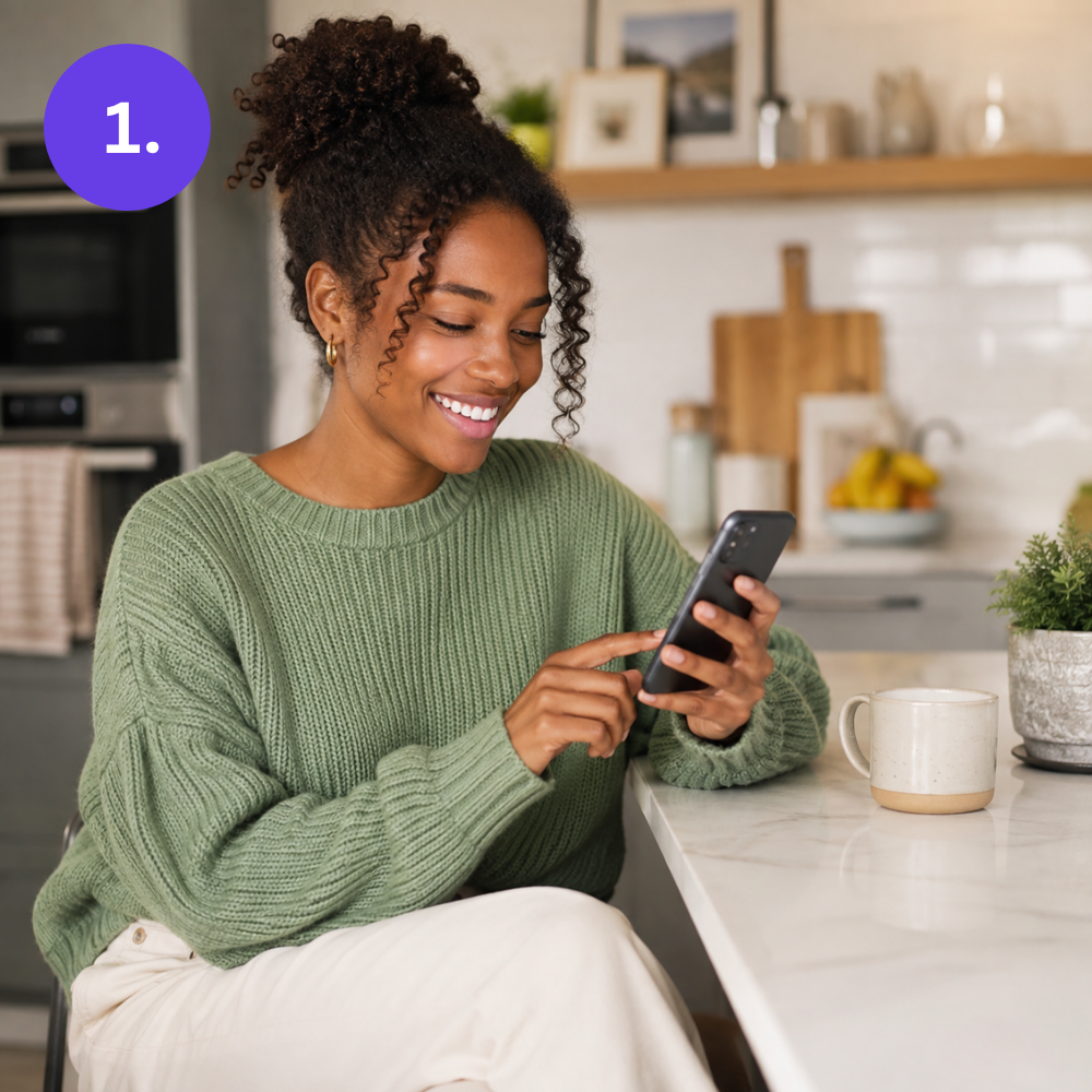 Woman smiling while browsing on her phone at a kitchen counter, signing up to list her car for sale